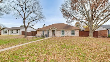A residential house with a brick exterior, a front yard with scattered leaves, and a large tree to the right.