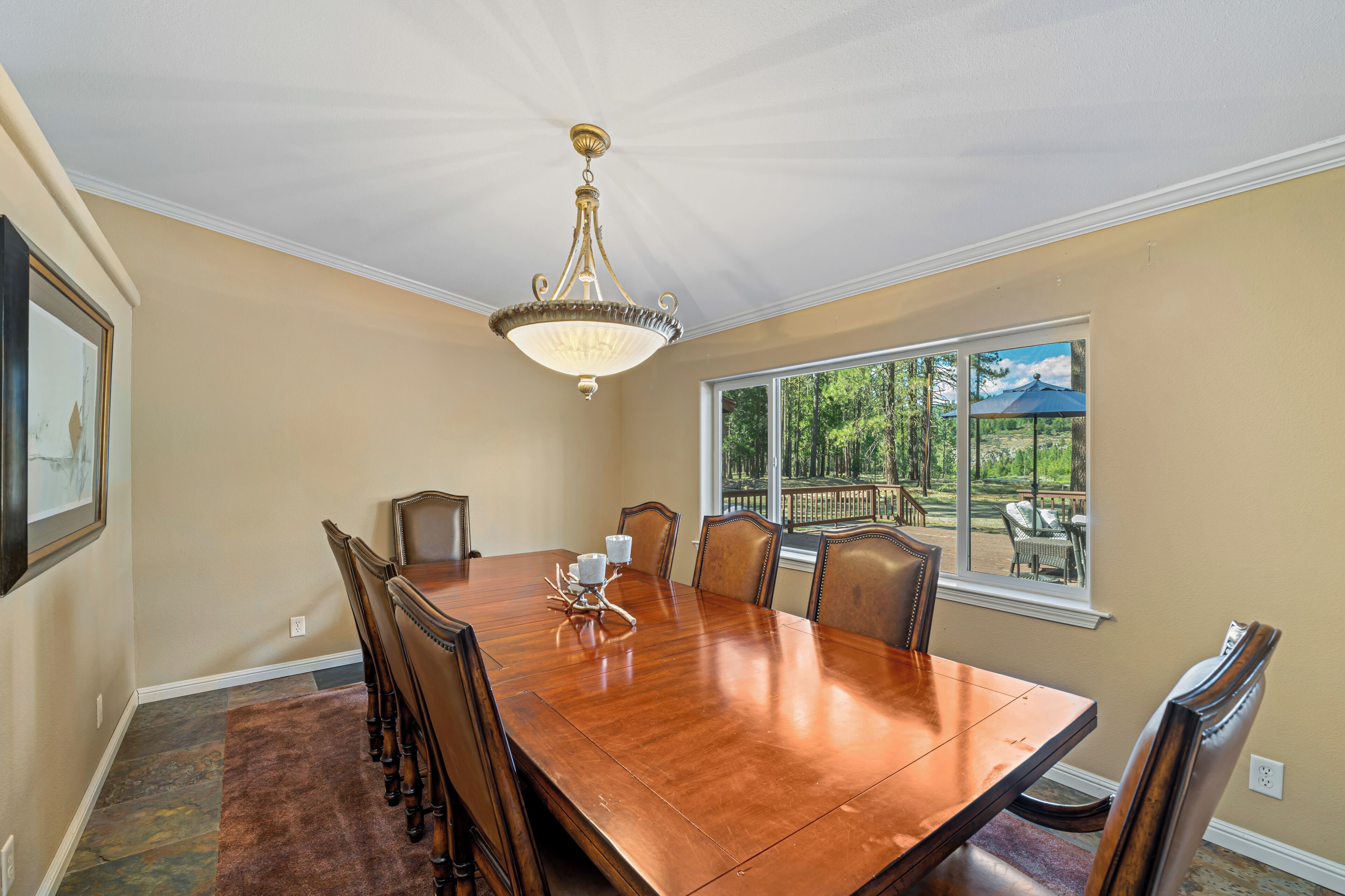 A dining room with a wooden table, chairs, a chandelier, and a window with a view of greenery.