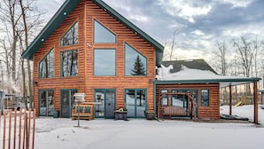 A log cabin with a large central window, a covered porch, and a snow-covered landscape. - Big Pine Lake Home w/ Dock & Game Room in Aitkin (Aitkin)