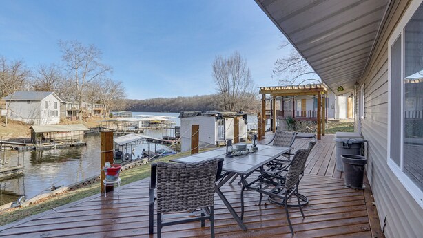 A wooden deck with outdoor furniture overlooking a lake, dock, and houses. - Lake Ozarks Home w/ Deck, Dock & Hot Tub (Versailles)