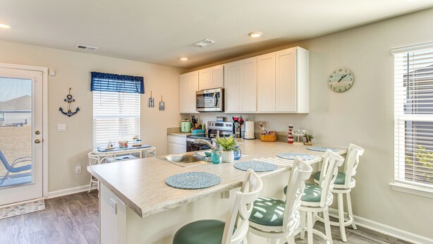 A modern kitchen with white cabinets, a central island with bar stools, a microwave, and a clock on the wall. - 3 Mi to Holden Beach! Family Home w/ Patio (Supply)