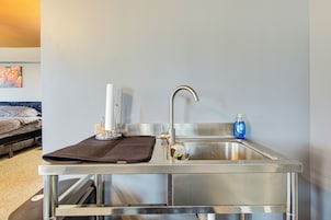 Un baño de hotel pequeño y funcional con un lavamanos de acero inoxidable, un espejo pequeño y una botella de jabón para manos.