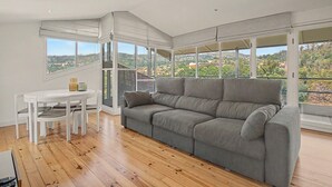 A spacious balcony with a grey sofa and white dining table, large windows offering a view of a hillside town. - Cantinho dos Avós – Country House with Mountain View, Wi-Fi and Air Conditioning (Sever do Vouga)