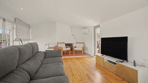 A modern living room with a grey sofa, wooden flooring, a flat-screen TV, and a dining area with chairs. - Cantinho dos Avós – Country House with Mountain View, Wi-Fi and Air Conditioning (Sever do Vouga)