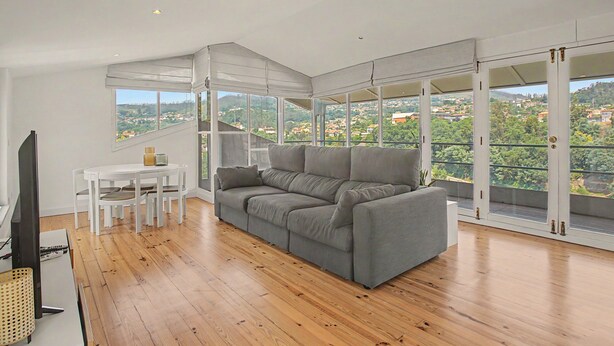 A modern living room with a grey sofa, white table, and chairs, large windows with a view of a town. - Cantinho dos Avós – Country House with Mountain View, Wi-Fi and Air Conditioning (Sever do Vouga)