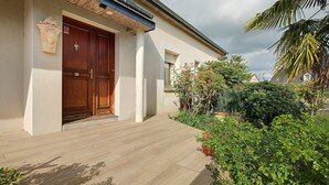 Une maison avec une porte d’entrée en bois, une façade blanche et un jardin où poussent différentes espèces de plantes.