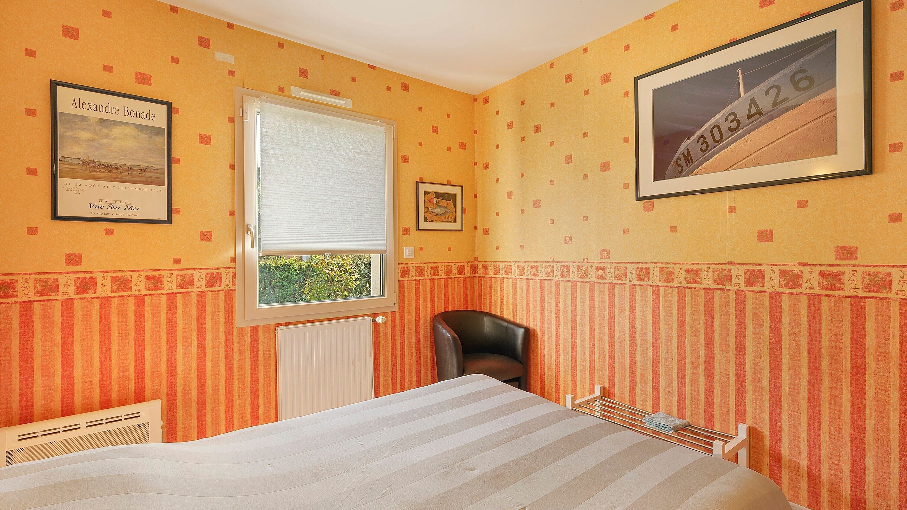 Une pièce avec un lit, une fenêtre, un radiateur, une chaise et des tableaux encadrés accrochés aux murs.