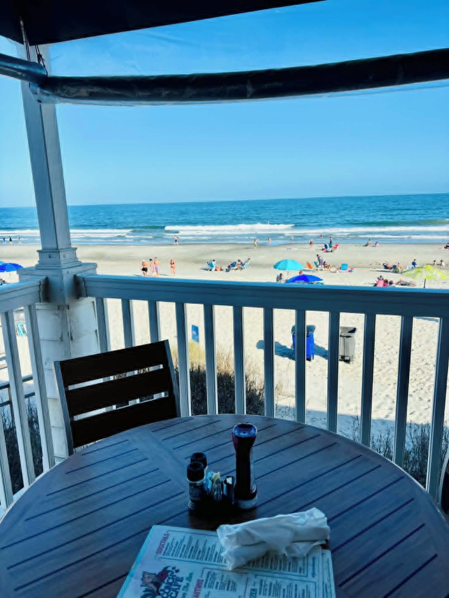 Een overdekt terras met uitzicht op het strand, een tafel met een krant en een fles.