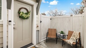 A front door with a wreath, a porch with two wicker chairs and a small table, and a wooden fence in the background.