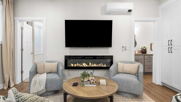 A modern living room with a flat-screen TV, a fireplace, two armchairs, a coffee table, and a kitchen area visible through an open door.