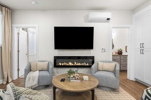 A modern living room with a flat-screen TV, a fireplace, two armchairs, a coffee table, and a kitchen area visible through an open door. - Cozy 1BR Boise Retreat | Central Location (Boise)