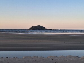 Una playa con una pequeña isla a lo lejos, agua tranquila y cielo despejado.