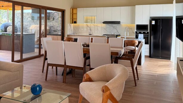 Amplia sala de estar con techo alto y vigas a la vista, comedor con mobiliario moderno y cocina con alacenas blancas y electrodomésticos de acero inoxidable.