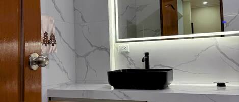 Un baño con pared de mármol, un lavamanos tipo cuenco negro, un cajón de madera y un espejo con marco iluminado.