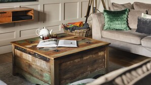 A rustic wooden coffee table with an open book and a teapot, a wicker basket with colorful blankets, a tripod floor lamp, and a beige sofa with green and gray pillows. - 1 Force Mill Barn in Satterthwaite (Ulverston)