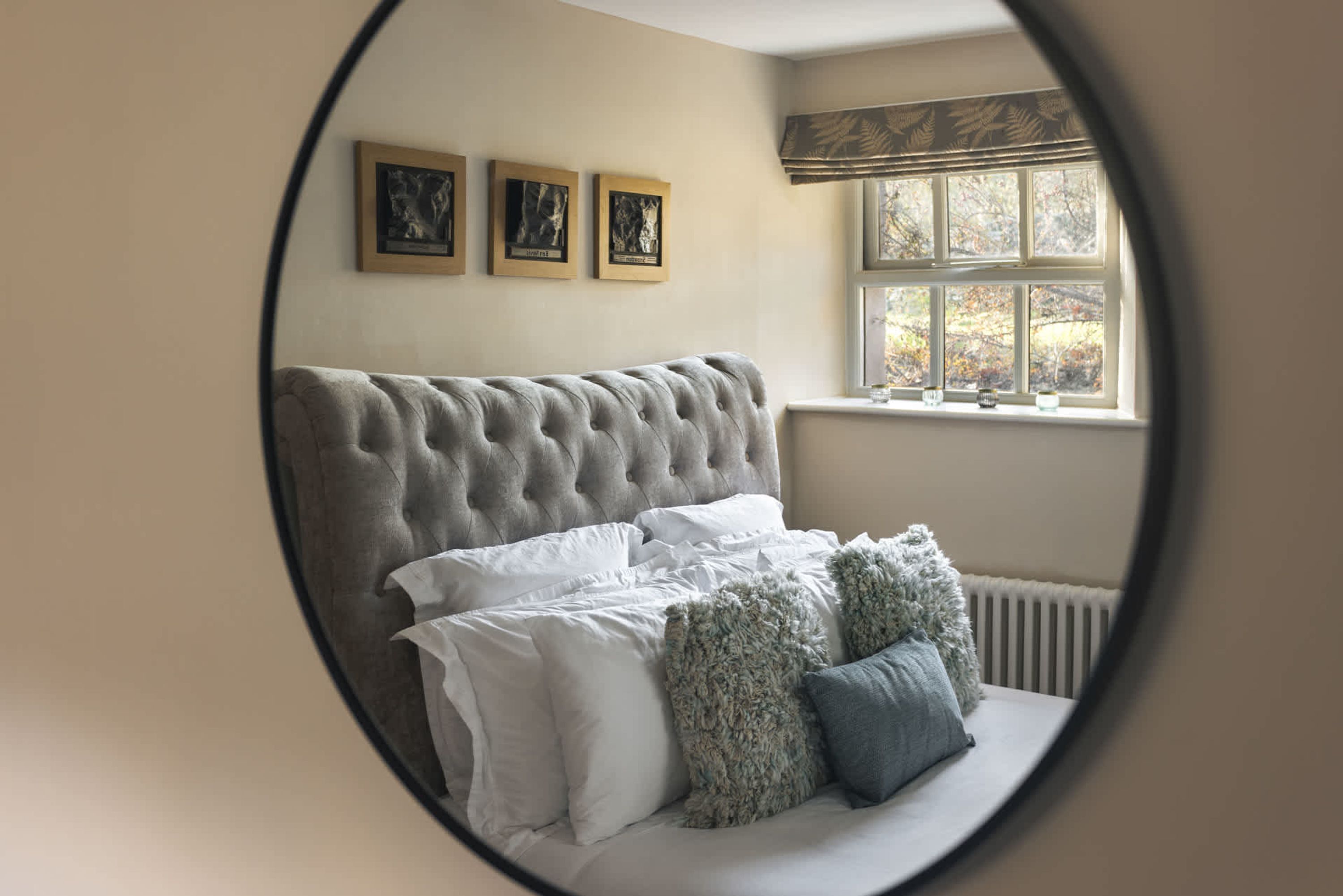 Un dormitorio con cabecera capitonada, una cama con ropa blanca y dos almohadas decorativas, una ventana con vista a árboles y tres cuadros en la pared.