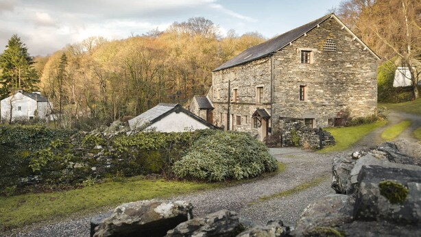 A stone building with a sloped roof, surrounded by a gravel path and greenery. - 1 Force Mill Barn in Satterthwaite (Ulverston)