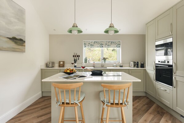 A kitchen with a central island, two wooden bar stools, pendant lights, and a window with a view of greenery. - The Lookout With 1 Bedrooms and 1 Bathrooms (Grange-over-Sands)