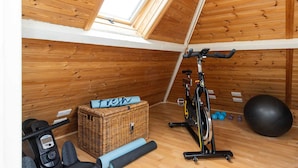 A small, wooden-paneled room with a sloped ceiling, a stationary bike, a basket with a towel, and exercise equipment. - Rural House With Swimming Pool and Gardens (Chichester)