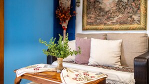 A room with a blue wall, a wooden table with a vase of green plants, a patterned tablecloth, a beige sofa with a decorative throw, and a large tapestry on the wall. - Petalidi Sunny Home One - Charming Garden Nest (Messini)