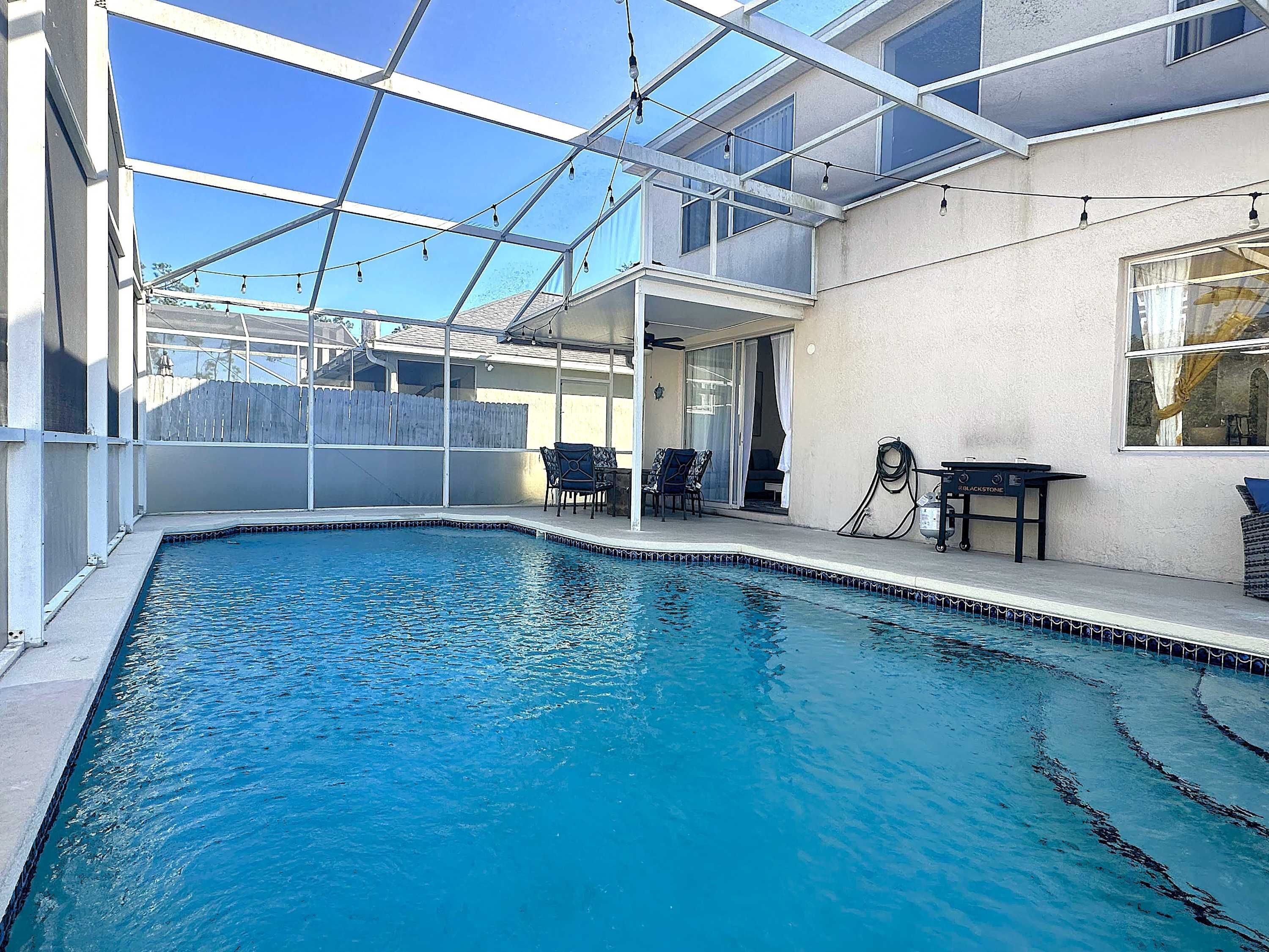 An indoor swimming pool with clear blue water, surrounded by a glass enclosure, and furnished with chairs and a table.