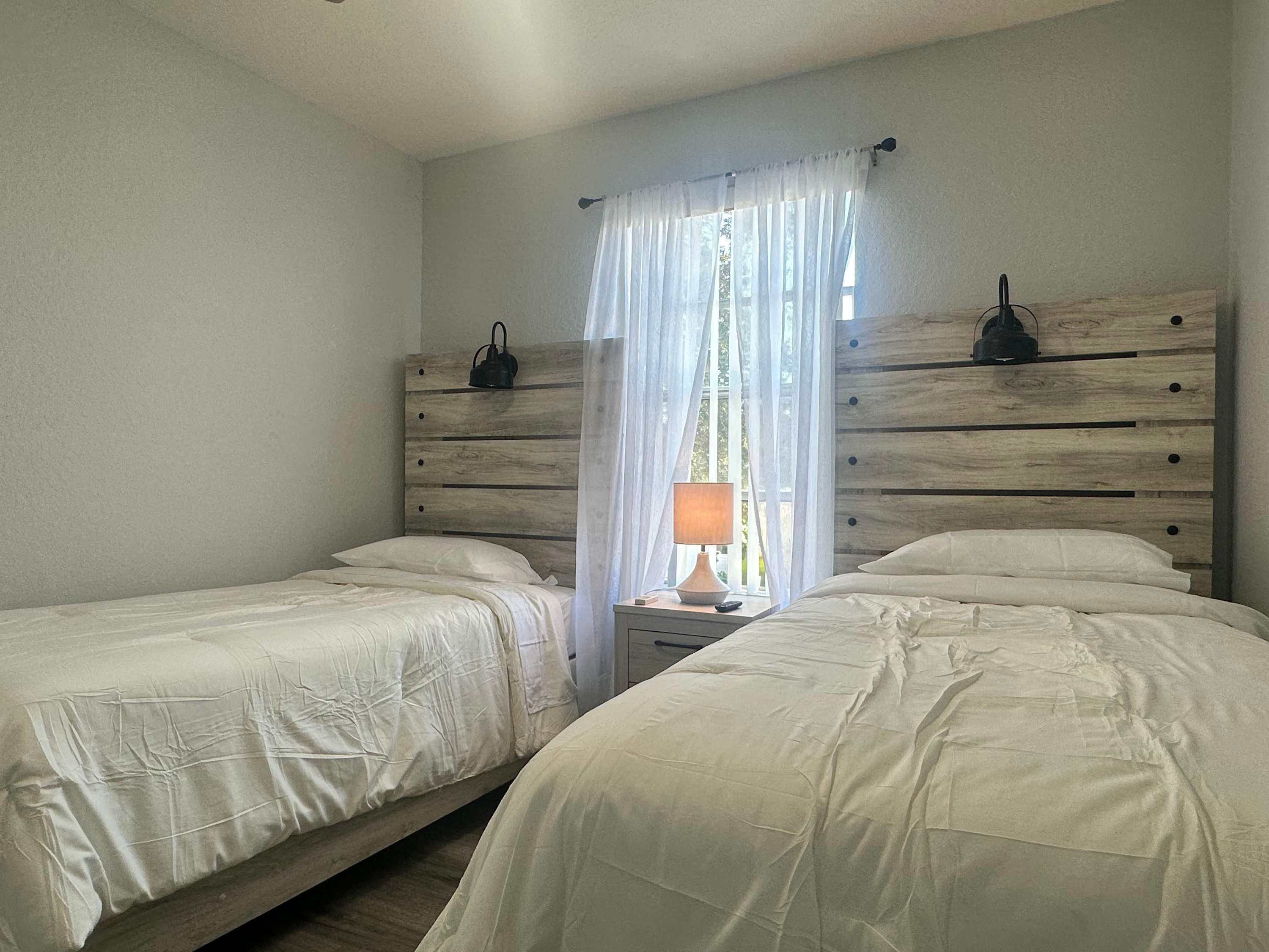 Une chambre avec deux lits, une table de chevet avec une lampe et une fenêtre avec des rideaux légers.