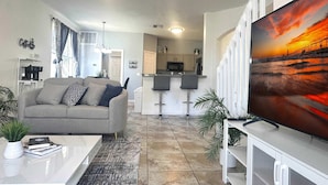 Una sala moderna con sofá, mesa de centro y televisor. Cuenta con una zona de cocina con taburetes altos y una gran pantalla de televisión.