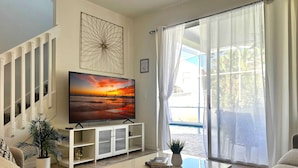 Una sala de estar con un televisor de pantalla plana grande sobre un soporte blanco, un sofá, una mesa de centro y una escalera con barandillas blancas.