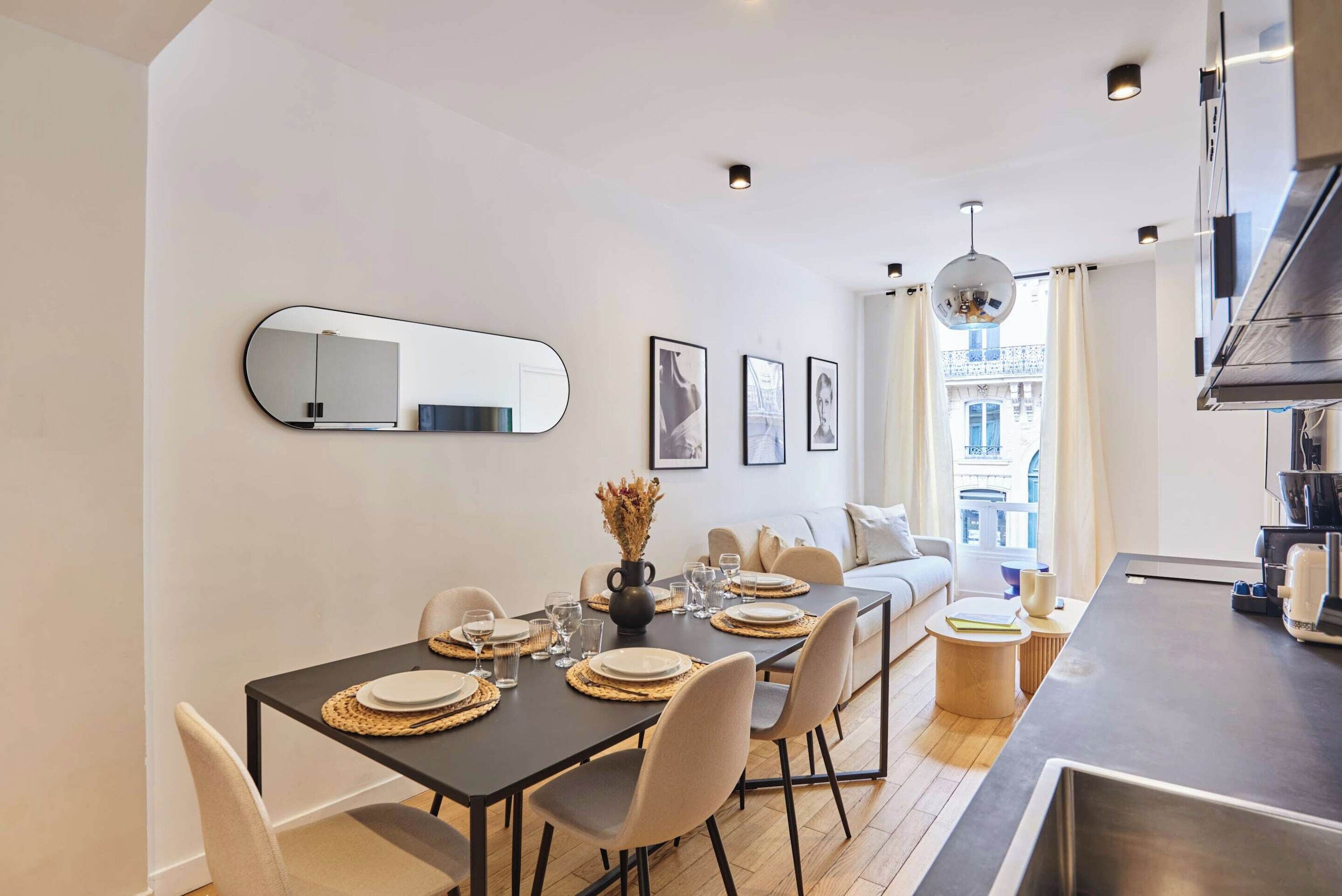 A modern dining area with a table set for four, a sofa, and a kitchen area with a large window.