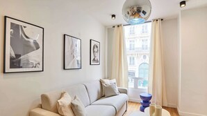 A modern living room with a sofa, framed artwork, a coffee table, and a view of a building through a window. - Sublim Apartment Paris Center Beaubourg Le Marais (Paris)