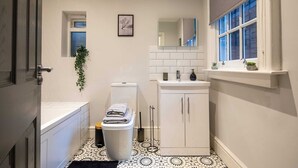 Ein kleines Badezimmer mit Toilette, Waschbecken und Spiegel. Ein Fenster mit Jalousien, eine kleine Pflanze und ein schwarz-weiß gemusterter Boden sind ebenfalls vorhanden.