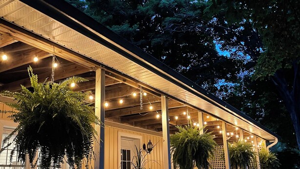 A covered porch with hanging plants and string lights, wooden railings, and a view of the surrounding trees. - Forest’s Edge Elora | Private Yard, Fire Pit, Near Downtown (Salem)