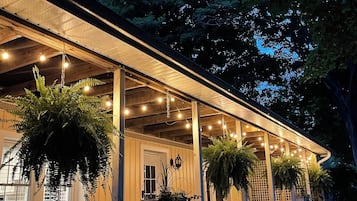 A covered porch with hanging plants and string lights, wooden railings, and a view of the surrounding trees.