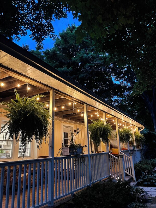 A covered porch with hanging plants and string lights, wooden railings, and a view of the surrounding trees.