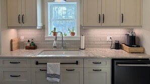 A kitchen with light-colored cabinets, a window with plants, a sink, and a coffee machine. - Forest’s Edge Elora | Private Yard, Fire Pit, Near Downtown (Salem)