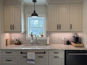 A kitchen with light-colored cabinets, a window with plants, a sink, and a coffee machine.
