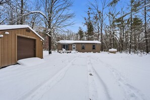 A wooden garage with a snow-covered roof, a small house with a front door and windows, and a snow-covered driveway leading to the house. - Lewiston Landing: Cozy Lake Access Getaway 3 Bedroom Home (Lewiston)