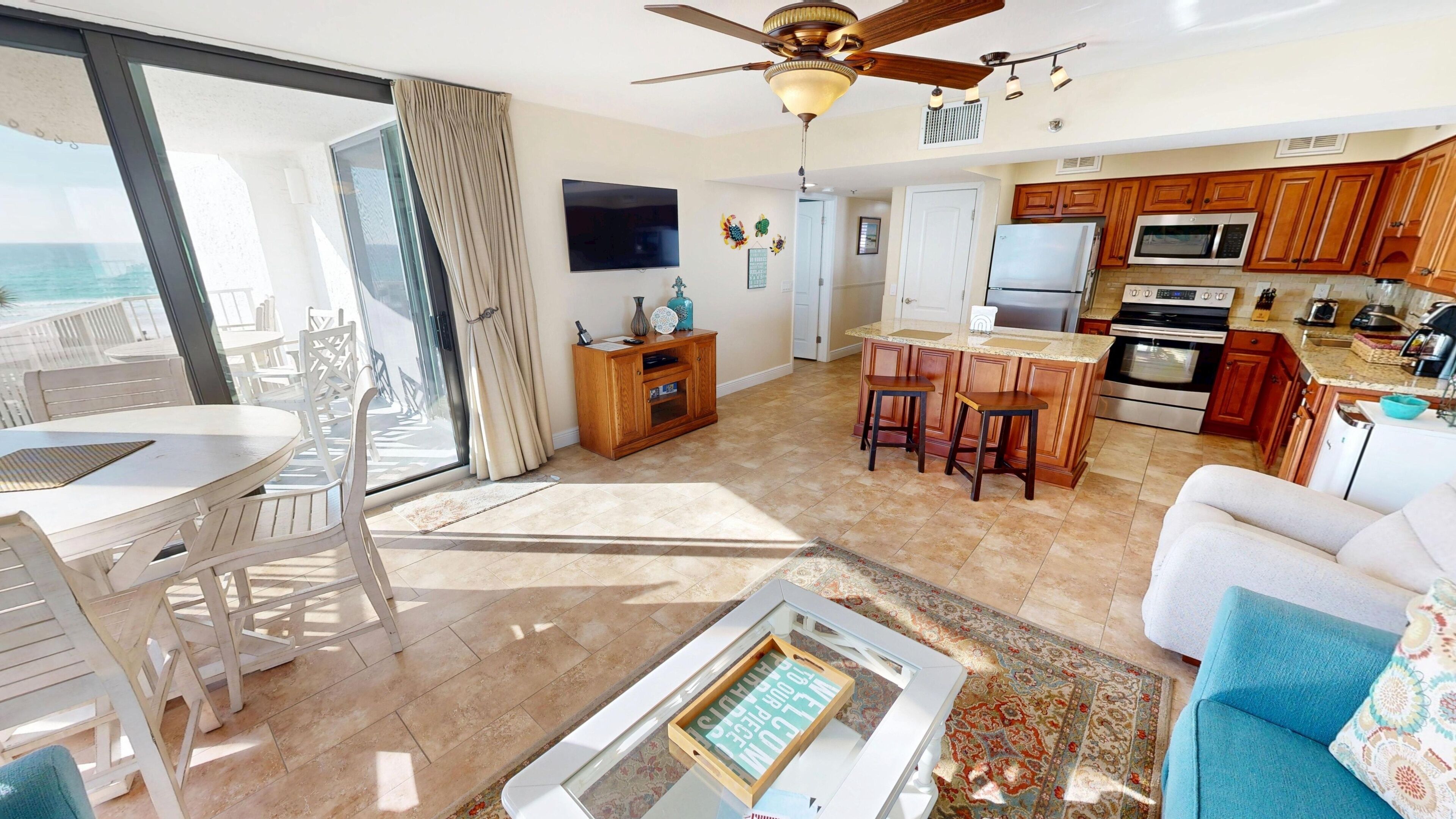 A spacious living area with a large sliding glass door leading to a balcony, a dining area with a table and chairs, a kitchen with wooden cabinets and modern appliances, and a ceiling fan.
