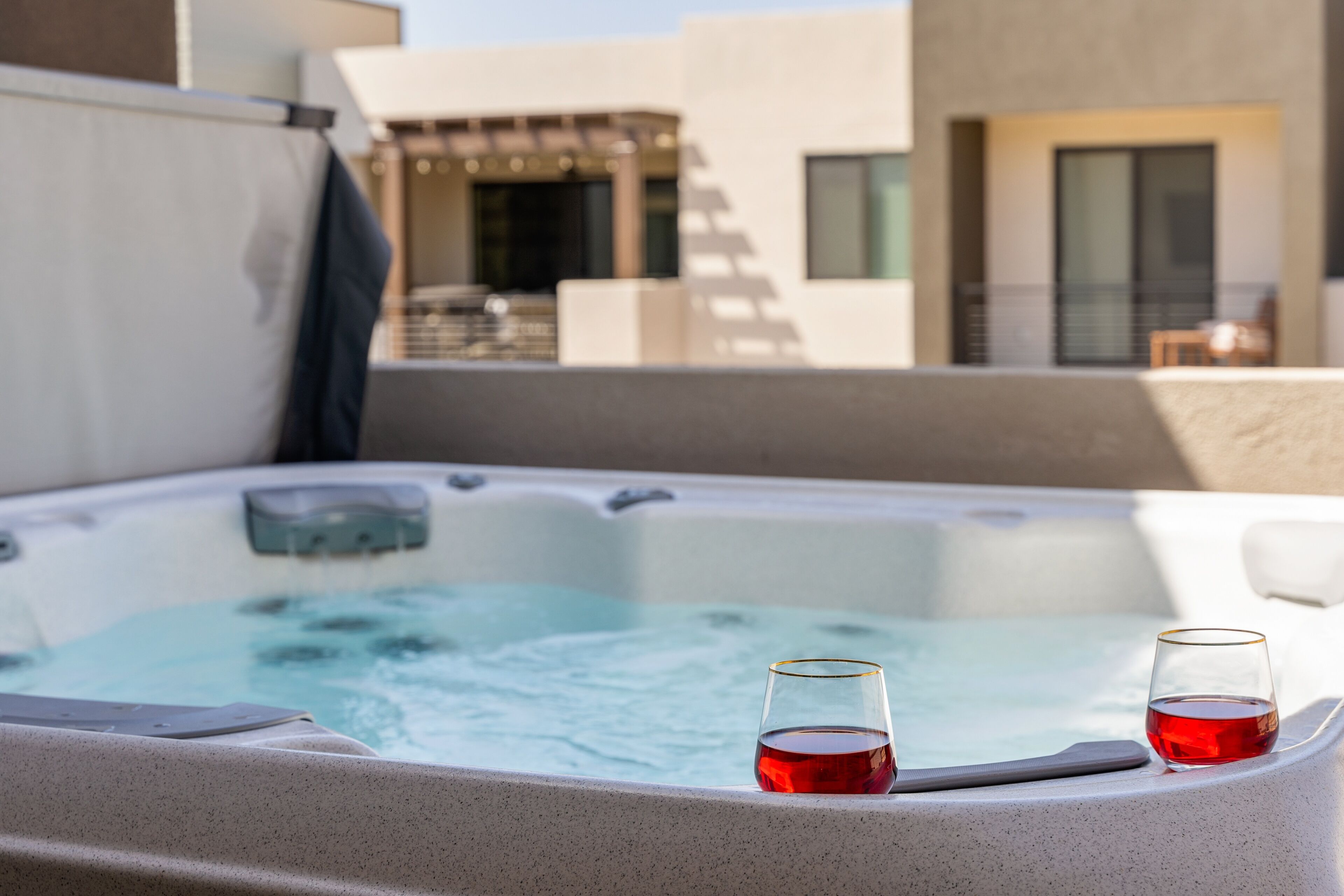 A hot tub with two glasses of red liquid, a white chair, and a building in the background.