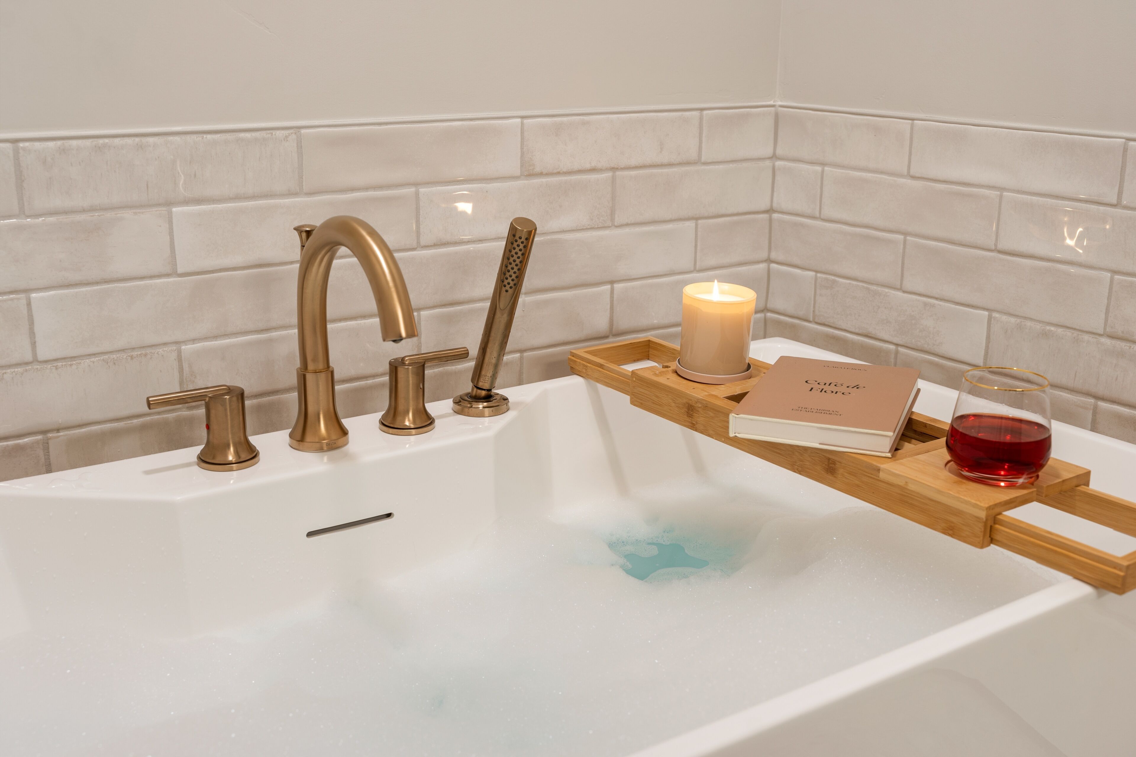 A bathroom with a white bathtub, gold faucets, a candle, and a tray with a book and glass.