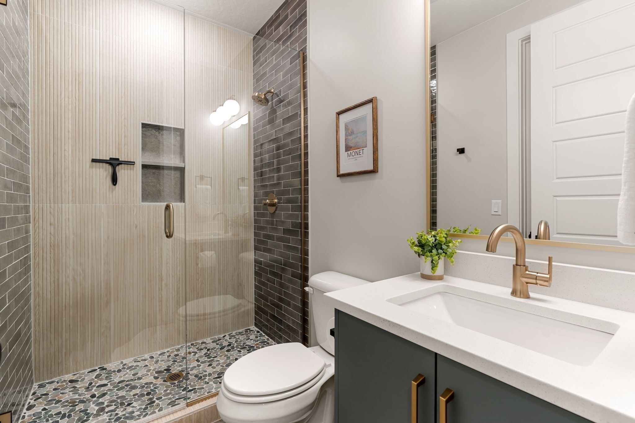 A modern bathroom with a glass-enclosed shower, a white toilet, a sink with a gold faucet, and a framed picture on the wall.