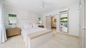 Une chambre spacieuse avec un grand lit, des tables de chevet, un ventilateur de plafond et une vue sur une piscine, visible à travers une porte ouverte.