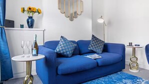 A blue sofa with patterned cushions, a small round table with a bottle and glasses, a decorative wall piece, and a floor lamp. - The Navy Nest Luxury Home Nottingham City Centre (Nottingham)
