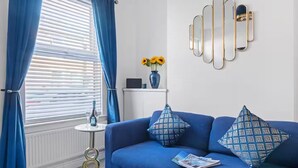 A room with a blue sofa, a small side table with a bottle and glasses, a vase with sunflowers, and a wall-mounted mirror. - The Navy Nest Luxury Home Nottingham City Centre (Nottingham)