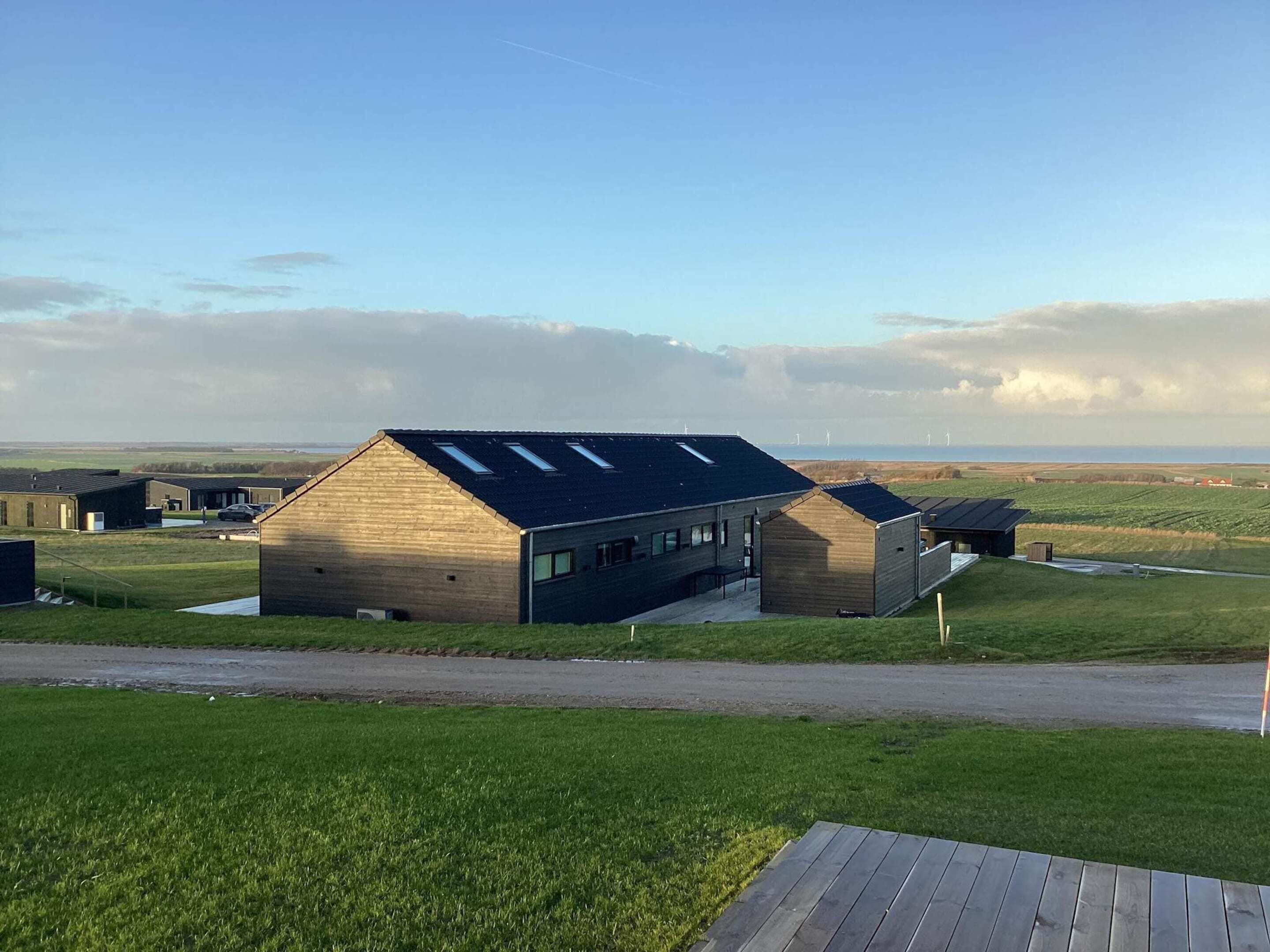 A modern building with a sloped roof and skylights, surrounded by green fields and a clear sky.