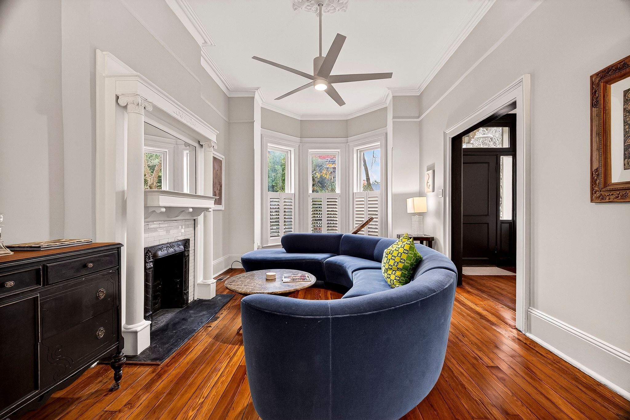A living room with a dark blue sofa, a fireplace, a ceiling fan, and a view of the outdoors through large windows.