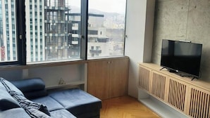 Una sala de estar moderna con un sofá seccional grande, un televisor sobre una consola de madera, y vistas urbanas a través de amplios ventanales.