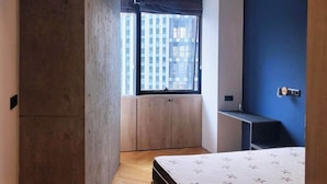 Un dormitorio moderno con una cama grande, almacenamiento integrado, una ventana con persianas y vista a la ciudad.