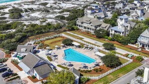 An aerial view of a coastal residential area with a large swimming pool surrounded by a white fence, a beach, and houses with a mix of architectural styles. - Gorgeous 7 Bedroom 7 Bath Large Home with Carriage House in Gated WaterSound! (WaterSound)