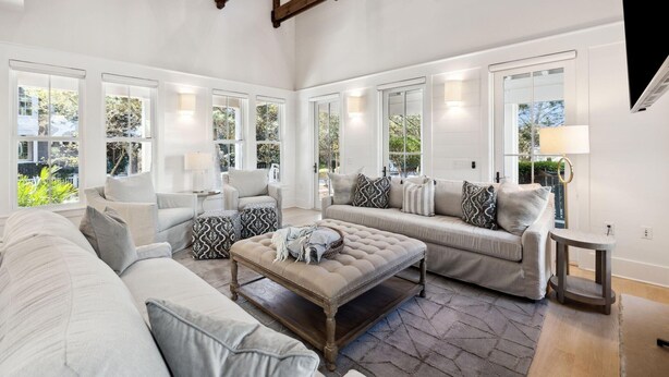 A spacious living room with a high ceiling, large windows, and a cozy seating area with a sofa and ottoman. - Gorgeous 7 Bedroom 7 Bath Large Home with Carriage House in Gated WaterSound! (WaterSound)